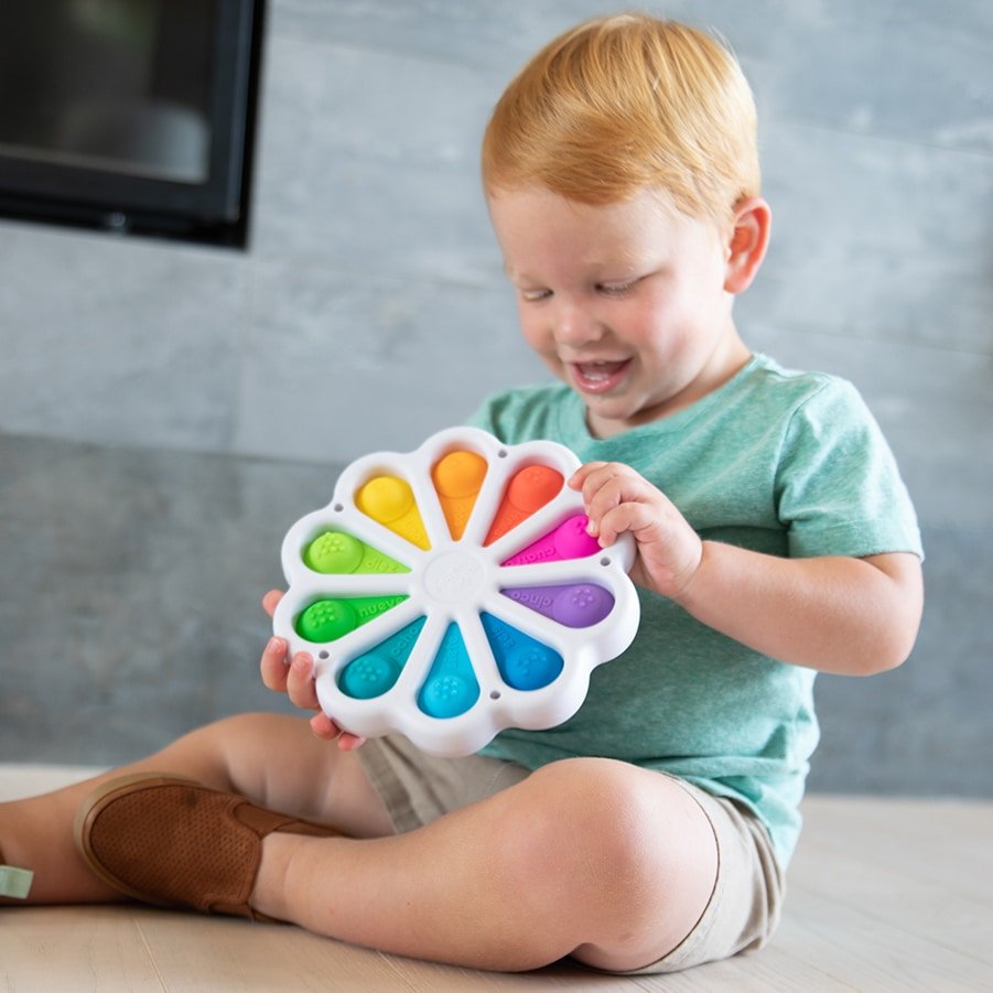 Rainbow flower-shaped pop toy with colorful wedge buttons, held by a smiling child.